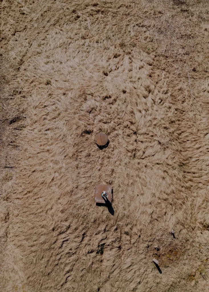 Top-down aerial view of a person standing on a square metal platform in a field of dry swirling grass, with a circular cover nearby